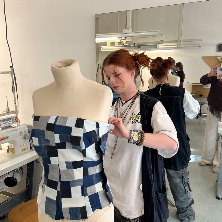 Una persona appunta un top di jeans patchwork su un manichino da sarto in un laboratorio di cucito, con una macchina da cucire, uno specchio e altre persone sullo sfondo.