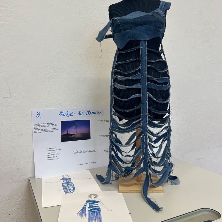 Un manichino espone un abito fatto di strisce di jeans tagliate e sovrapposte, simile a una gabbia toracica. Accanto, ci sono schizzi di stilisti di moda e una bacheca di progetto intitolata "Libertà degli elementi" con testo e l'immagine di un tramonto.