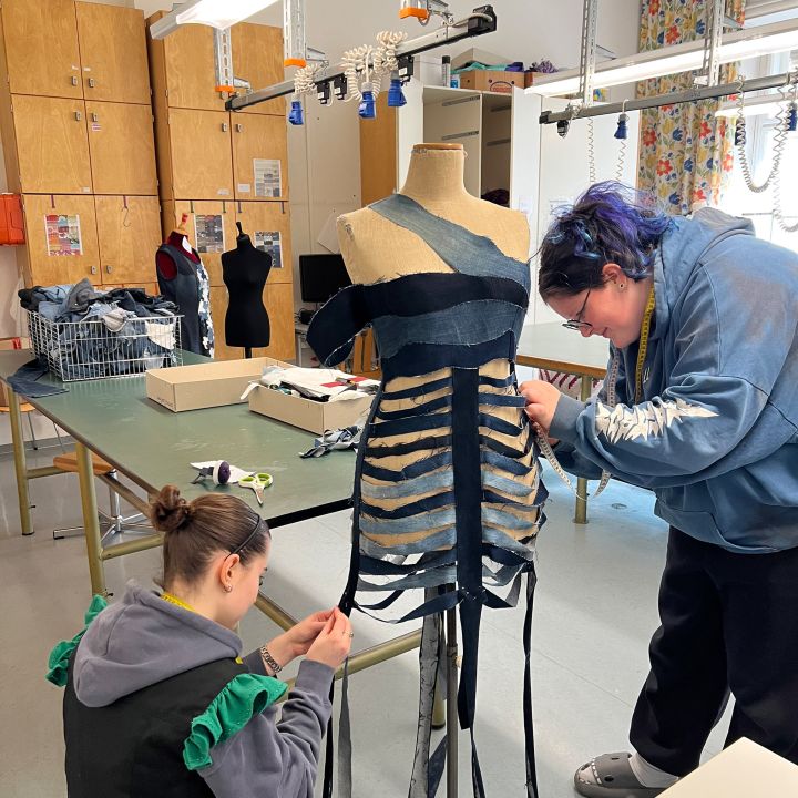 Due persone lavorano insieme in un'aula su una forma per abiti, applicando strisce di tessuto scuro per creare un capo unico. La stanza è arredata con tavoli da lavoro, manichini, armadi in legno e tende colorate sullo sfondo.
