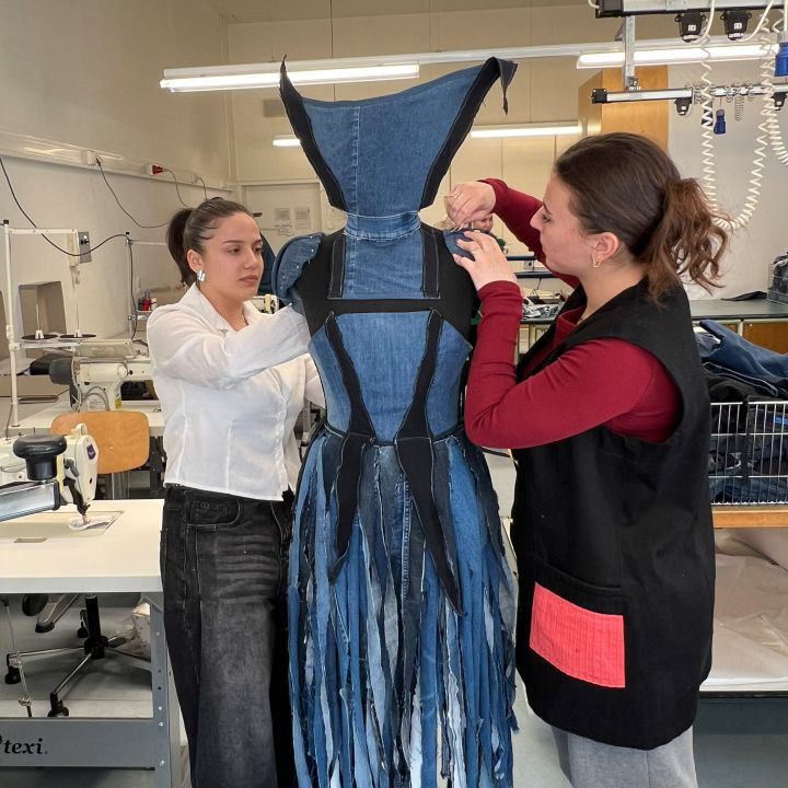 Due donne lavorano insieme su un modello di abito, sistemando un abito di jeans blu destrutturato con frange. Si trovano in un luminoso atelier di cucito, dove si intravedono macchine da cucire e strumenti sullo sfondo.