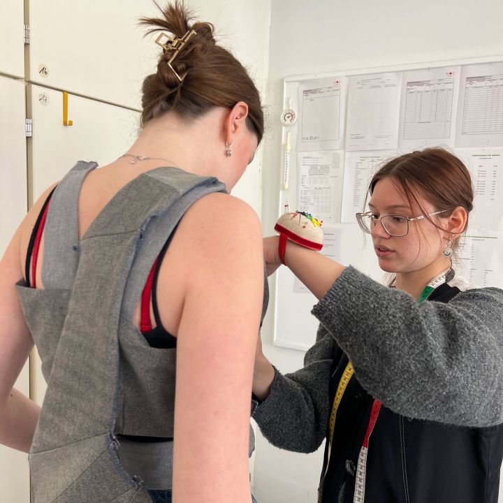 Due donne si trovano in una sartoria; una indossa un capo parzialmente confezionato, mentre l'altra lo sta sistemando con strumenti da cucito e un metro da sarta intorno al collo. Sul muro sullo sfondo si possono vedere dei cartamodelli.