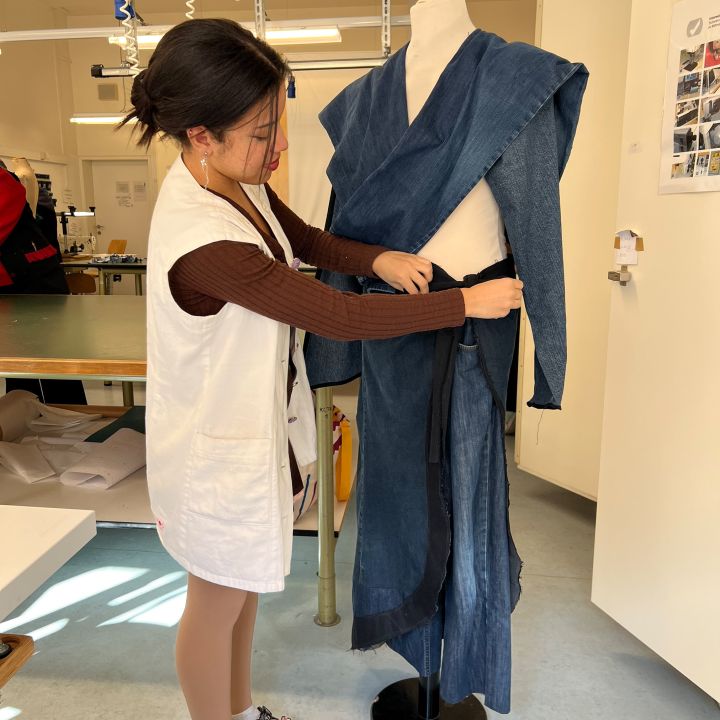 Una persona che indossa un top marrone, un gilet bianco e sandali sta sistemando un indumento blu simile a un kimono su un manichino in uno spazio di lavoro ben illuminato.