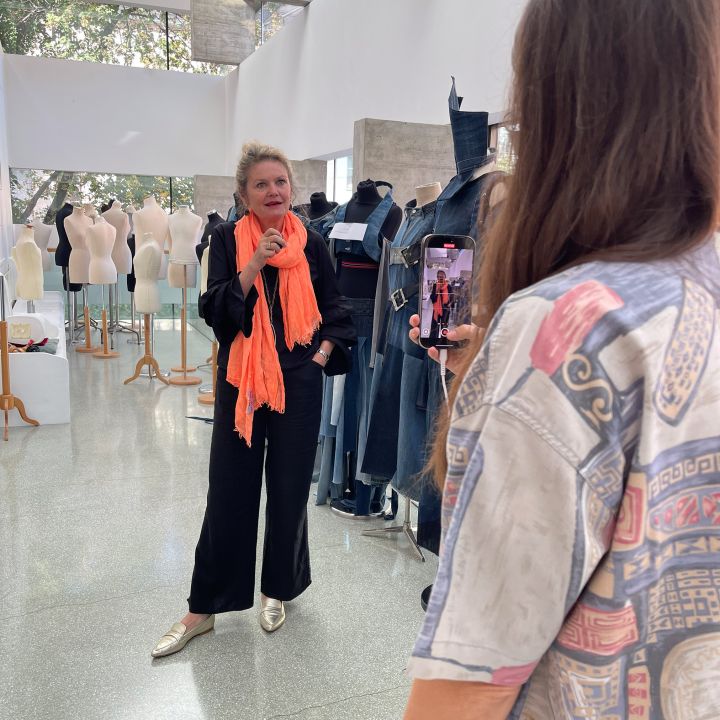 Una donna in abito nero e sciarpa arancione brillante posa tra manichini e forme di abbigliamento, mentre un'altra persona la fotografa con uno smartphone in uno studio luminoso e moderno.