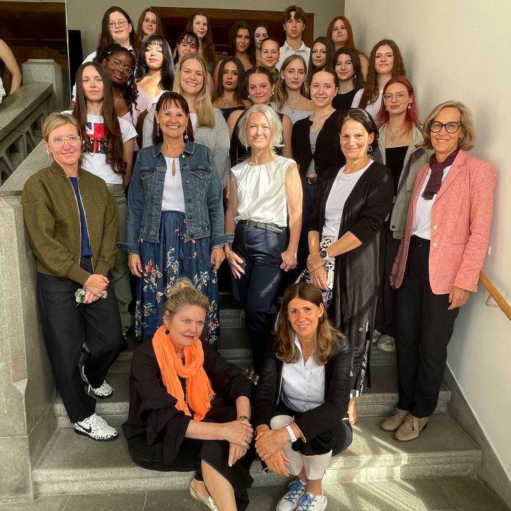 Un gruppo di donne e ragazze posa insieme su una scalinata, sorridendo alla fotocamera. Alcune sono in piedi, altre sedute sui gradini. L'ambiente sembra essere un interno con luce naturale.