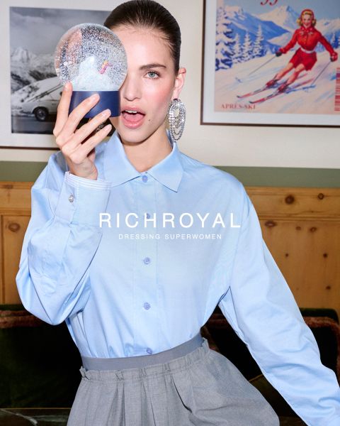 Una donna in camicia azzurra e pantaloni grigi si tiene una palla di neve davanti a un occhio. Posa in una stanza con pareti in legno e un poster di sci, e indossa grandi orecchini. Il testo recita "RICHROYAL DRESSING SUPERWOMEN".