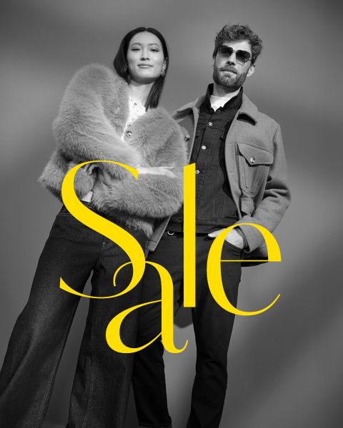 Una donna elegante con un cappotto di pelliccia e un uomo con occhiali da sole e giacca posano con sicurezza. La foto in bianco e nero è sovrapposta dalla scritta gialla in grassetto "Saldi".
