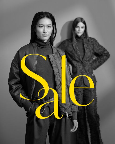 Due donne posano in eleganti abiti scuri davanti a uno sfondo grigio con la parola "Sale" in grandi lettere gialle che attraversano l'immagine.