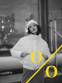 Una persona con un piumino bianco e un cappello posa all'aperto vicino a un edificio; una linea diagonale gialla attraversa l'immagine con due lettere "O" gialle su entrambi i lati. La foto è in bianco e nero.