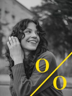 Una donna con i capelli ricci sorride e guarda di lato. Indossa un maglione ed è seduta all'aperto. Un grande simbolo di percentuale giallo si sovrappone alla foto in bianco e nero.