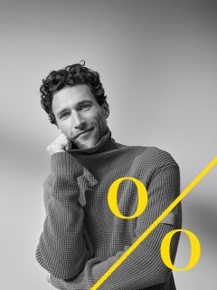 Un uomo con i capelli ricci e la barba indossa un grosso dolcevita e appoggia il viso sulla mano sorridendo. Un grande simbolo di percentuale giallo è sovrapposto diagonalmente all'immagine.