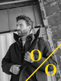 Un uomo con la barba è in piedi davanti a uno sfondo industriale e si sistema la giacca. La foto in bianco e nero mostra grandi simboli di percentuale gialli e una linea diagonale gialla nell'angolo inferiore destro.