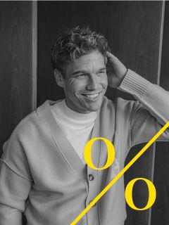 Un uomo con i capelli ricci che indossa un cardigan chiaro e una camicia bianca è seduto sorridendo e tiene una mano dietro la testa. L'immagine in bianco e nero mostra due grandi "o" gialle e una linea diagonale gialla sul bordo inferiore.