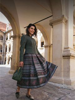 Una donna con una gonna fantasia e una giacca verde è in piedi sotto archi di pietra, sorride e tiene in mano una borsa verde.