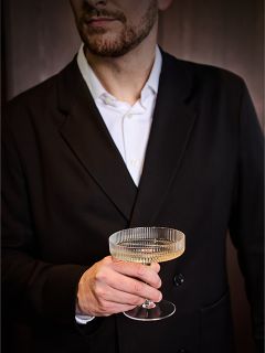 Un uomo in abito nero e camicia bianca tiene in mano un bicchiere da champagne trasparente e rigato, con il viso parzialmente fuori dall'inquadratura.