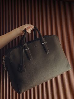 Una mano tiene una grande cartella di pelle nera con due manici davanti a uno sfondo marrone ondulato.