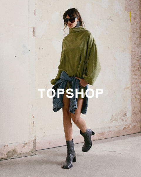 Una donna con un top verde oversize, una giacca di jeans legata in vita, stivali neri e occhiali da sole è in piedi davanti a un muro consumato. Il testo "TOPSHOP" è sovrapposto.