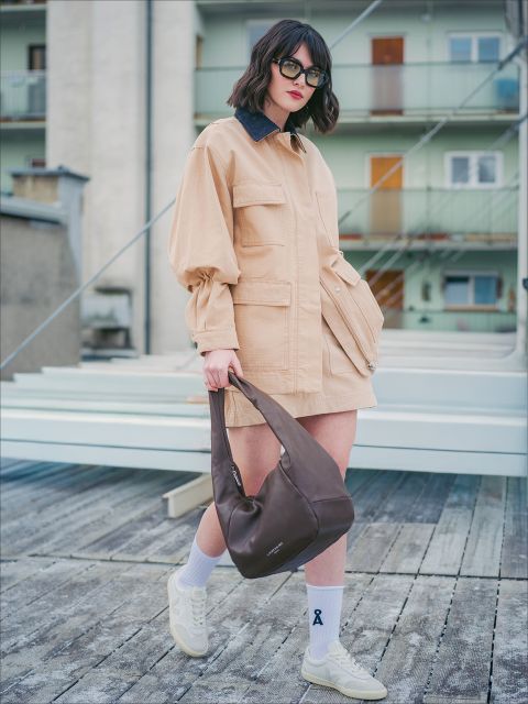 Una donna con giacca e gonna beige, occhiali da sole e scarpe da ginnastica bianche indossa una borsa marrone mentre si trova all'aperto su una terrazza di legno.