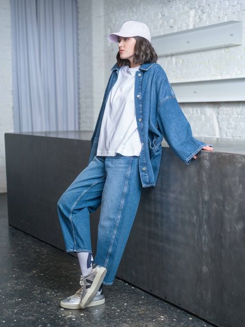 Una persona che indossa una giacca di jeans, una t-shirt bianca e un cappello bianco è appoggiata a un bancone scuro in un interno moderno.