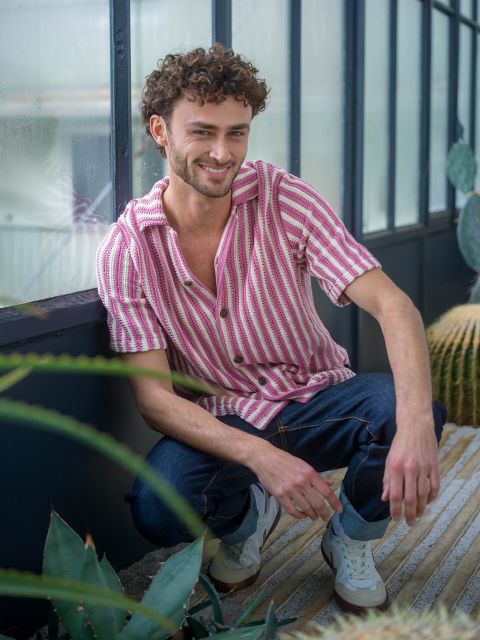 Un uomo con i capelli ricci, con una camicia a righe rosa e jeans, è accovacciato in una casa vicino a delle piante e sorride alla telecamera.