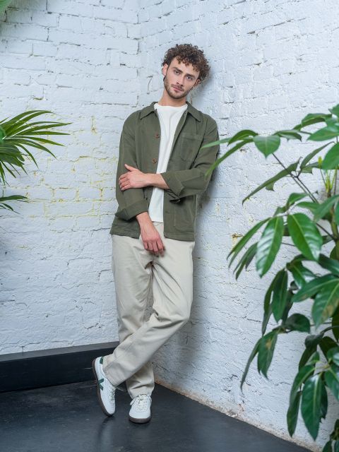 Un uomo con i capelli ricci, con una giacca verde, una camicia bianca e pantaloni beige, è appoggiato a un muro di mattoni bianchi, circondato da piante verdi.