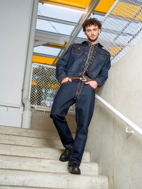 Un uomo con giacca di jeans scura e jeans è in piedi su una scala, con un piede più in alto, e si tiene al corrimano, dietro di lui una recinzione metallica e un lucernario giallo.