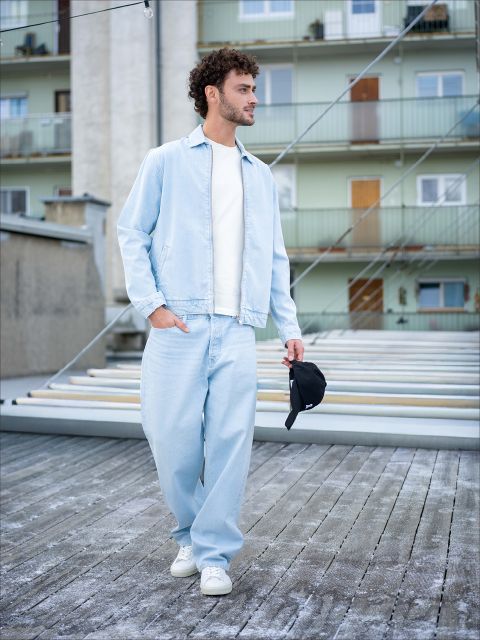 Un uomo con jeans azzurri, giacca abbinata e scarpe da ginnastica bianche cammina all'aperto e tiene in mano un berretto nero, sullo sfondo si vedono case residenziali.