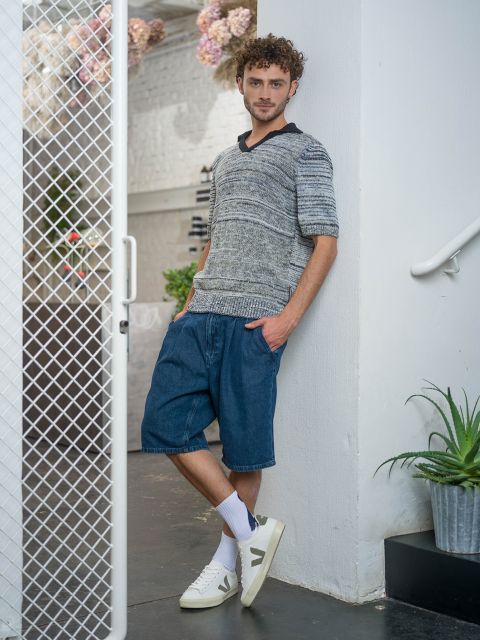 Un uomo è in piedi accanto a un muro bianco, indossa un maglione grigio di lana, pantaloncini di jeans, calzini bianchi e scarpe da ginnastica, con un piede incrociato sull'altro.