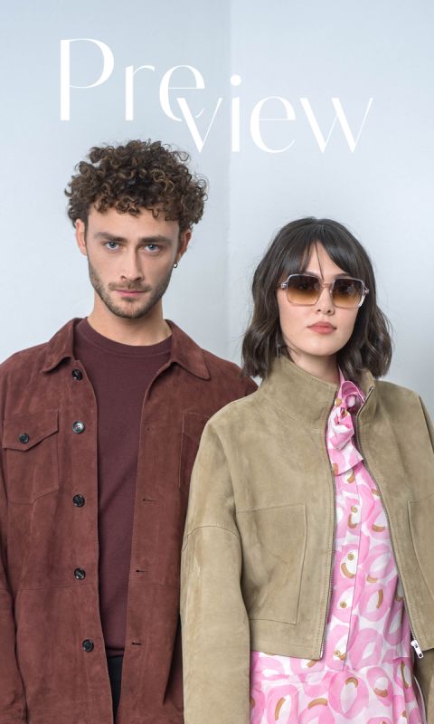 Un uomo con una giacca marrone e una donna con occhiali da sole e una giacca marrone chiaro sopra un abito rosa fantasia sono in piedi uno accanto all'altra davanti a un semplice sfondo chiaro.