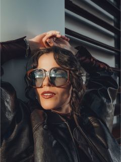 Una donna con grandi occhiali da sole e una giacca di pelle nera si appoggia all'indietro con le mani sopra la testa, la luce del sole si riflette nei suoi occhiali.
