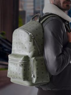 Una persona con una giacca nera indossa un grande zaino MCM verde oliva strutturato con tasche con cerniera e si trova all'aperto in un ambiente urbano.