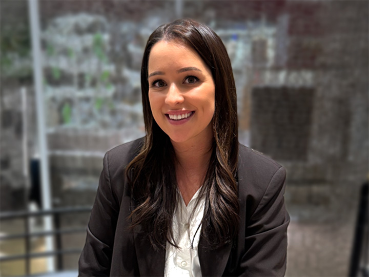 Una donna con lunghi capelli scuri in un blazer nero e una camicia bianca sorride mentre è seduta in una casa, con uno sfondo sfocato.