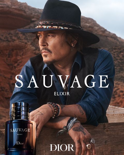 Un uomo con cappello e gilet è appoggiato a una ringhiera di legno nel deserto, in primo piano un flacone del profumo Sauvage Elixir Dior con logo.