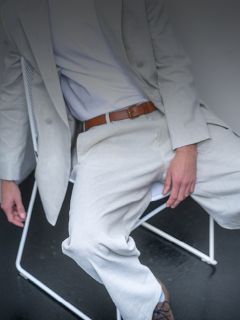 Una persona in un abito grigio chiaro e una camicia bianca è seduta su una sedia bianca, si vede una cintura marrone e il suo viso non è visibile.