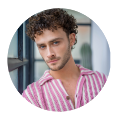 Uomo con capelli castani ricci e occhi azzurri, che indossa una camicia a righe rosa e bianche e guarda la telecamera in un interno vicino a una finestra.