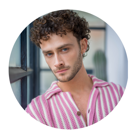 Uomo con capelli castani ricci e occhi azzurri, che indossa una camicia a righe rosa e bianche e guarda la telecamera in un interno vicino a una finestra.