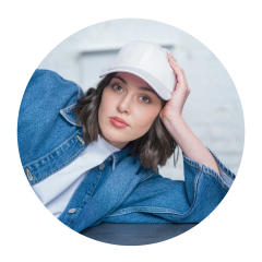 Donna con cappello bianco e giacca di jeans, appoggiata sul braccio e che guarda la fotocamera con sguardo neutro.