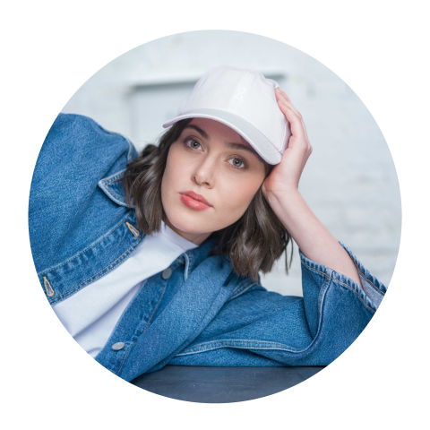 Donna con cappello bianco e giacca di jeans, appoggiata sul braccio e che guarda la fotocamera con sguardo neutro.