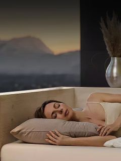 Una donna dorme su un fianco nel letto con un cuscino beige, accanto a una finestra che mostra un paesaggio montano sfocato al crepuscolo.