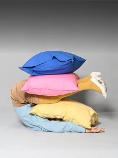 Una persona è rannicchiata sul pavimento, coperta da tre grandi cuscini colorati, da cui spuntano solo le sue gambe vestite di giallo e le scarpe bianche.