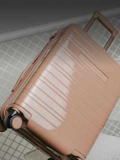 Una valigia rigida lucida, marrone chiaro, con ruote e maniglia estraibile è appoggiata su un pavimento piastrellato.