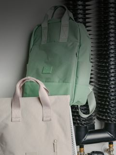 Uno zaino verde menta e una borsa tote beige sono appesi a una parete davanti a tubi e bobine neri.