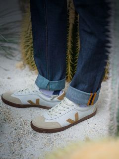 Persona in jeans scuri con risvolto e scarpe da ginnastica bianche con accenti marrone chiaro, in piedi su un terreno sabbioso vicino a piante di cactus verdi.