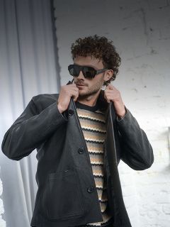 Uomo con capelli ricci che indossa occhiali da sole, un maglione a righe e una giacca di pelle nera, che si sistema il colletto davanti a un muro di mattoni bianchi.