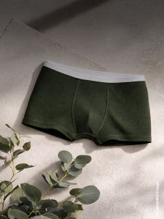 Boxer da uomo verde scuro con elastico bianco esposti su una superficie strutturata, con foglie di eucalipto nelle vicinanze in una luce soffusa e naturale.