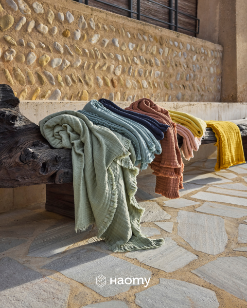 Copriletti colorati e strutturati drappeggiati su una rustica panca di legno all'aperto, con un muro di pietra e un marciapiede sullo sfondo. Logo Naomy in basso.