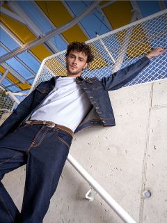 Un uomo con una maglietta bianca, una giacca di jeans scura e jeans è in piedi accanto a una ringhiera appoggiata a un muro di cemento e a una recinzione, sopra una tenda gialla.