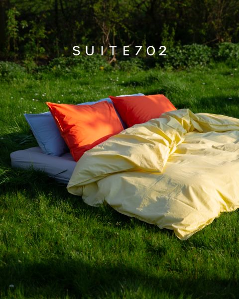 Un letto con cuscini arancioni e rossi brillanti e un copriletto giallo si trova all'aperto sull'erba verde alla luce del sole. Sopra c'è scritto "SUITE702".