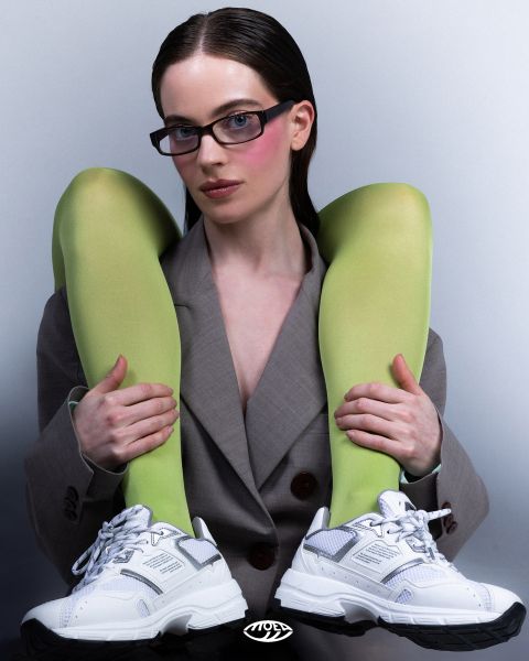 Una donna in un blazer grigio indossa occhiali e scarpe da ginnastica bianche e tiene le gambe con calze verde brillante sopra le spalle, davanti a uno sfondo semplice.