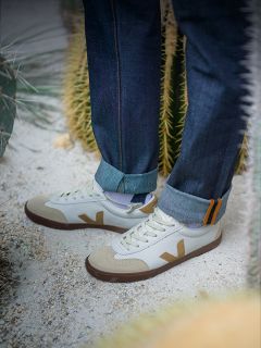 Persona in jeans arrotolati in piedi su un terreno sabbioso vicino a cactus, indossa scarpe da ginnastica bianche con accenti beige e marroni e un logo a "V" sul lato.