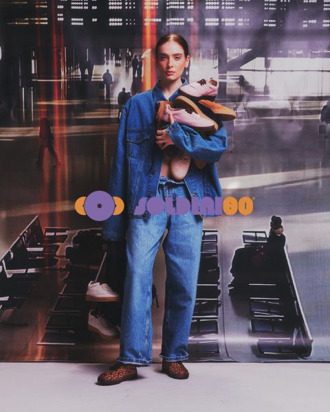 Una donna in giacca di jeans e jeans è in un terminal aeroportuale e tiene in braccio diverse paia di scarpe da ginnastica.